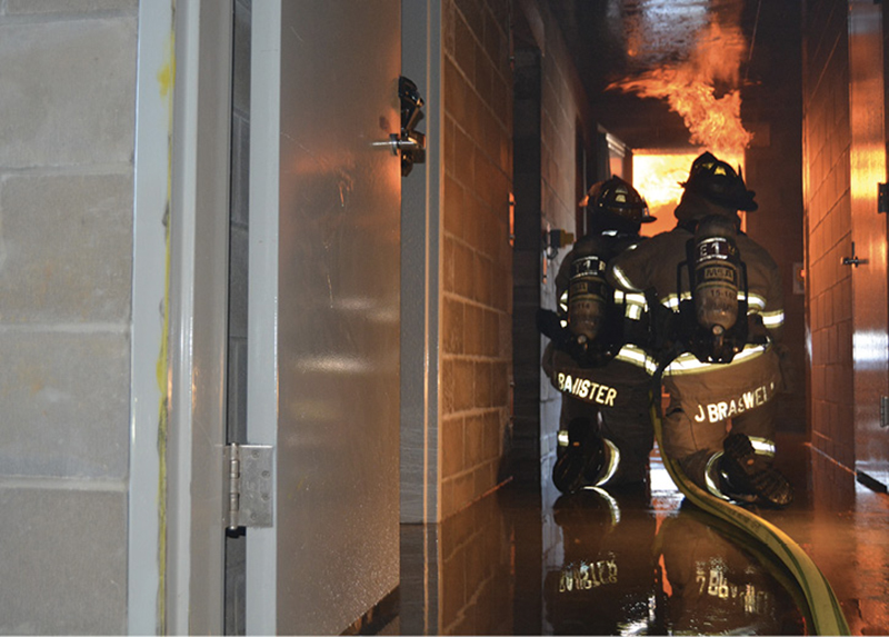 Firefighters advance a Key Hose Combat Ready attack line toward a live fire in a training building.